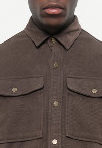 Cămașă maro cu nasturi, fabricată dintr-un material texturat, având două buzunare pe piept cu accesorii aurii și un design clasic al gulerului.