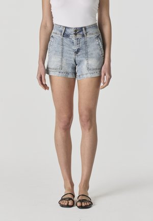 Mujer vistiendo shorts de mezclilla de tiro alto en azul claro con grandes bolsillos frontales, combinados con sandalias negras de tiras y una blusa blanca sin mangas.