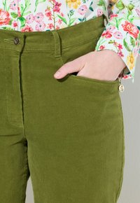 Olivgrüne Cordhosen mit einer Fronttasche, sichtbarem Reißverschluss und einem goldenen Anhängeral. Strukturiertes Finish, körperbetonter Schnitt.