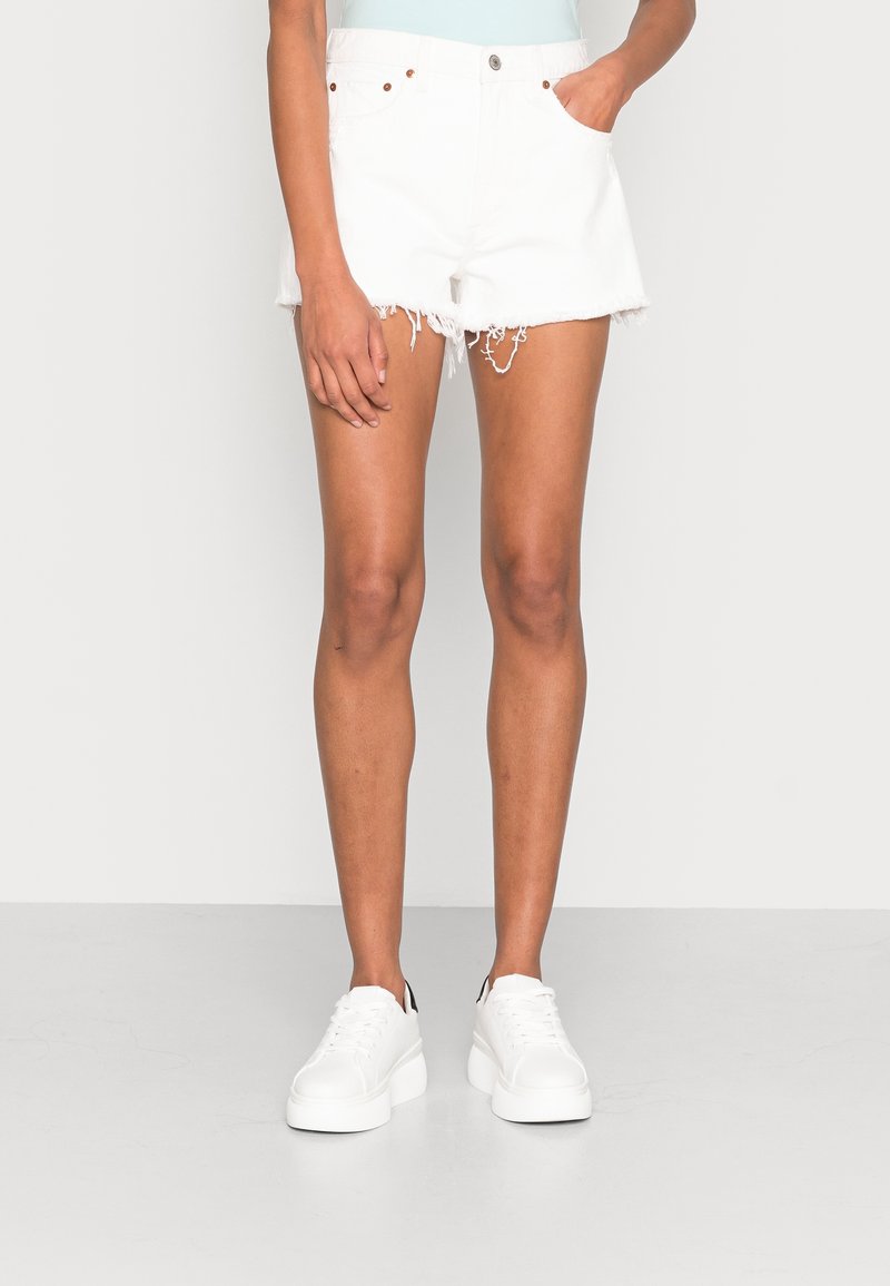 Shorts in denim bianco con orli sfrangiati, dotati di cinque tasche e rivetti in rame. Abbinati a sneakers bianche. Design minimalista, senza motivi.