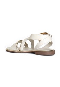 White leather sandal with cross straps and circular cutouts. Flat brown sole with gold accents on the edges. Back strap features a buckle.