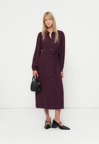 Rochie lungă, cu nasturi, dintr-un material de culoare purpuriu închis, cu talie legată. Prezintă mâneci lungi și o croială dreaptă, asortată cu accesorii negre.