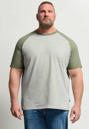 Mężczyzna o krótkich włosach i brodzie, ubrany w jasnoszarą koszulkę z oliwkowozielonymi rękawami raglanowymi i niebieskie dżinsy, stojący na jednolitym tle.