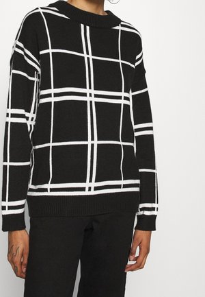 Persona che indossa un maglione nero con motivo a griglia bianca e pantaloni neri, in piedi contro uno sfondo chiaro uniforme.
