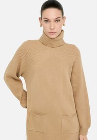 Maglione turtleneck beige a maglia con texture a coste, maniche lunghe, due tasche anteriori e cuciture delle spalle angolate per una vestibilità comoda.