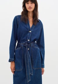 Denim haljina u tamno plavoj boji, s dugim rukavima, V-izrezom i srebrnim dugmetima. Uključuje vezani remen i prednje džepove.