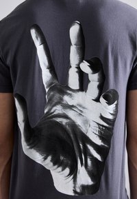 Črna bombažna majica s kratkimi rokavi, na hrbtni strani sledi velika, enobarvna podoba roke, ki dela znak za mir. Gladka tekstura.