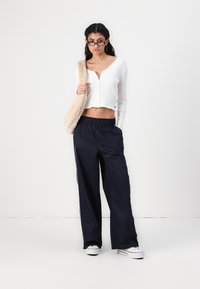 Weißes, geripptes Crop-Top mit langen Ärmeln, kombiniert mit einer navyblauen, weit geschnittenen Hose und weißen Sneakers, akzentuiert mit schwarzen seitlichen Streifen und einer Pelztasche.