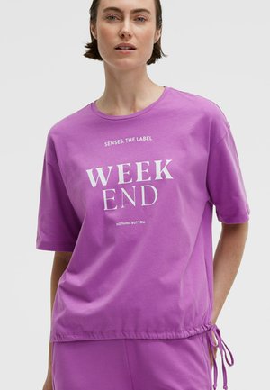 Woman wearing a purple short-sleeve shirt with "WEEK END" printed in white and matching purple pants with side drawstrings.