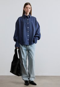 Επίκτητο πουκάμισο από σκούρο μπλε τζιν με κουμπιά μπροστά, κλασικό γιακά και ραφές ίδιου χρώματος, σε συνδυασμό με τζιν ανοιχτού μπλε και μαύρη τσάντα tote.