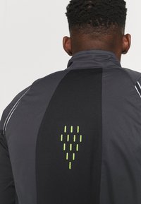 Veste de sport grise avec un panneau en maille noire et des accents réfléchissants. Détail notable : un motif géométrique vert au dos.