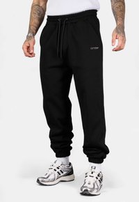 Schwarze Jogginghose aus weichem Stoff, mit elastischem Bund, Kordelzug und Bündchen an den Knöcheln. Gesticktes Logo auf der Vorderseite.