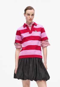 Polo shirt rayé rouge et rose avec un col et des manches retroussées, associé à une jupe noire ajustée. Logo notable sur la poitrine.