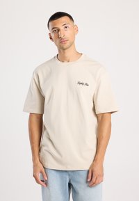 Beige puuvillane t-särk lühikeste varrukatega, ümar kaelus ja tikkpilt vasakul rinnal. Kombineeritud helesiniste teksadega.