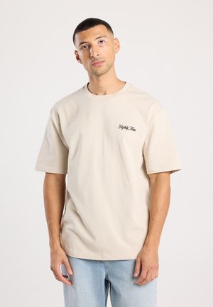 Beige katoenen t-shirt met korte mouwen, ronde hals en geborduurd logo op de linkerkant van de borst. Gepaard met lichtblauwe denim jeans.