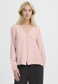 Blond naine, kes kannab heleroosa pluusi, millel on nupud ees ja musti pükse, seistes lihtsa heleda tausta ees.