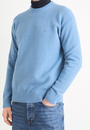 Hellblau gestrickter Pullover mit geripptem Saum und Bündchen, der ein dezentes Logo auf der Brust zeigt. Wird über einem dunklen Rollkragenpullover getragen.