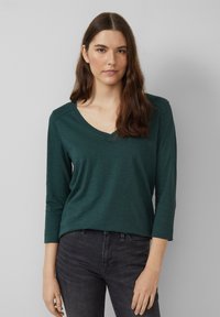Dunkelgrünes Langarmoberteil mit V-Ausschnitt, aus weichem Material gefertigt. Verfügt über eine lockere Passform mit Nahtdetails an den Schultern. Kombiniert mit dunkler Jeans.