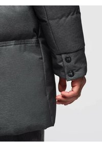 Cappotto puffer grigio con una finitura testurizzata, in primo piano un polsino con bottoni e una mano parzialmente visibile. Il materiale sembra morbido e isolante.