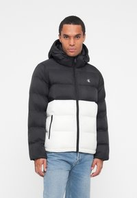 Schwarze und weiße Steppjacke mit Kapuze, ausgestattet mit einem Rei�ßverschluss, schwarzen Akzenten und einem Logo auf der Brust; glatte Textur und gestepptes Design.