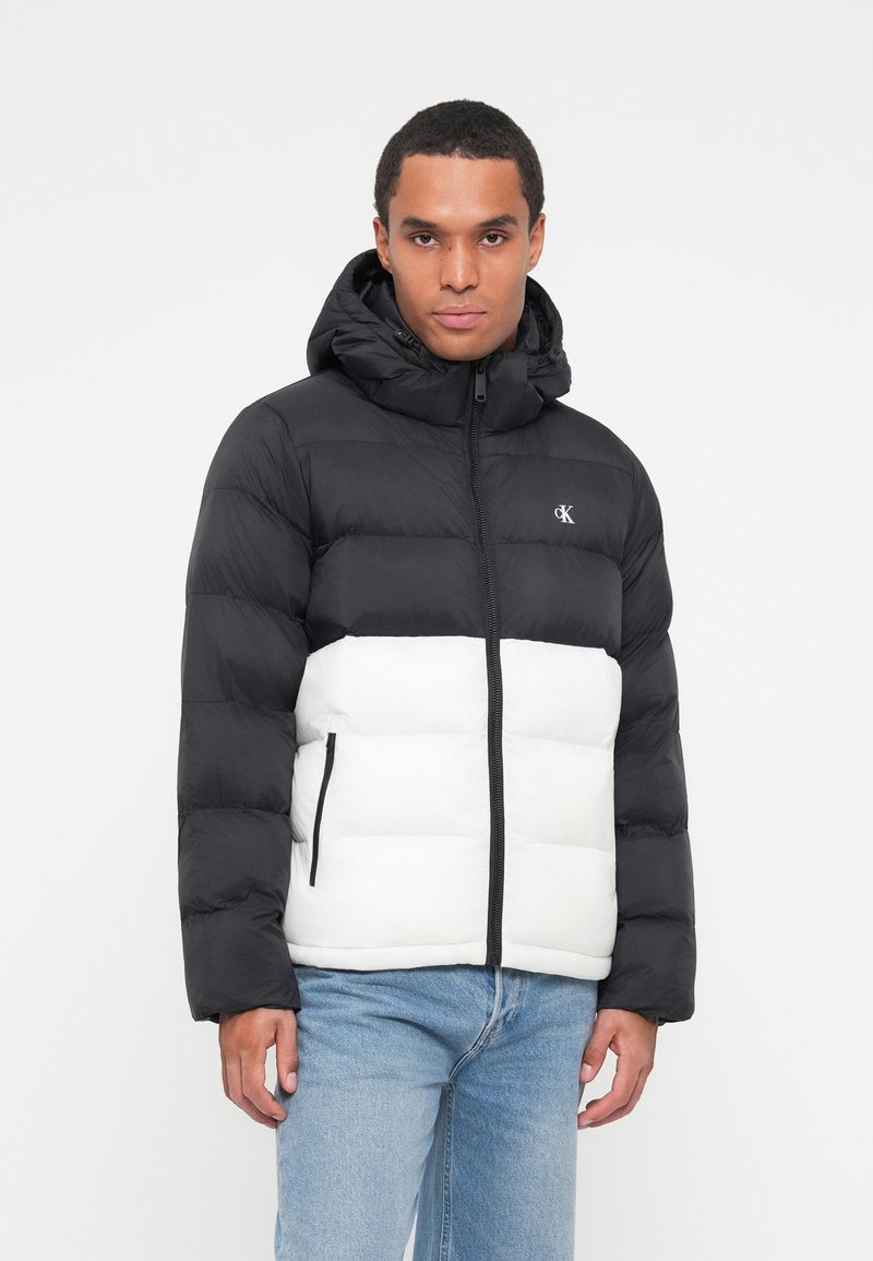 Schwarze und weiße Steppjacke mit Kapuze, ausgestattet mit einem Reißverschluss, schwarzen Akzenten und einem Logo auf der Brust; glatte Textur und gestepptes Design.