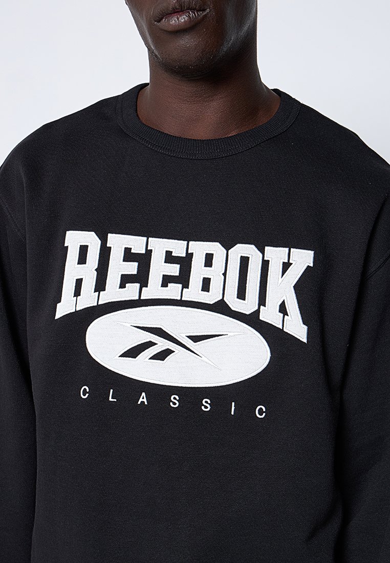 Uomo che indossa una felpa nera con un grande logo bianco "Reebok Classic" sul petto, su uno sfondo chiaro e semplice.