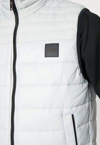 Ljust grå vadderad väst med horisontell sömnad, svart dragkedja och en fyrkantig svart läderlogotyp på bröstet, med sidfickor.