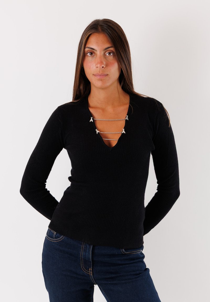 Top nero a maniche lunghe con costine, caratterizzato da un profondo scollo a V con dettagli in catena argentata e piccole decorazioni, abbinato a jeans blu scuro.