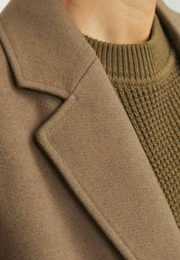 Abrigo de lana marrón con cuello pico, que presenta una textura suave, combinado con un suéter verde oliva de canalé debajo. Vista en primer plano.