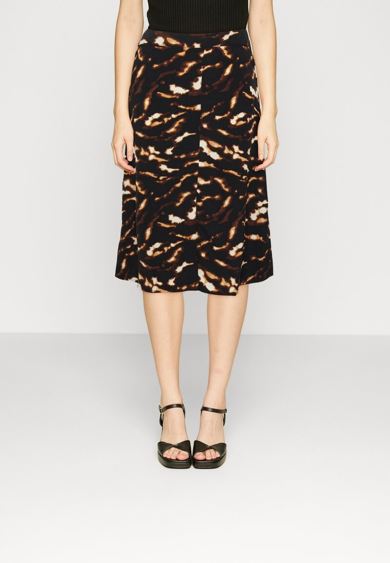 Femme portant une jupe mi-longue à motifs abstraits noirs et bruns, avec des sandales à talons ouvertes noires, debout devant un fond uni.