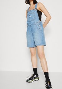 Salopette in denim azzurro chiaro con bretelle regolabili, tasca frontale e bottoni in argento. Abbinata a una canotta nera e scarpe sportive.