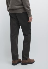 Homme portant un pantalon marron foncé, des chaussures en daim marron et un pull en maille gris, debout devant un fond clair uni.