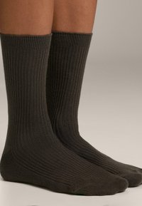 Gambe che indossano calze a costine scure, lunghe fino al polpaccio, in piedi su uno sfondo neutro.