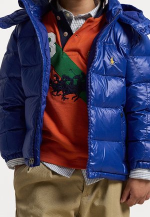 Enfant portant une doudoune bleue sur une chemise rayée boutonnée et un polo orange avec une bande diagonale verte et un logo cheval brodé.