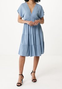 Svetle modré šaty do kolen s hlubokým V-výstřihem, krátkými rukávy a vrstvenou sukní. Vyrobené z měkké tkaniny s hladkým povrchem. Černé sandály na otevřenou špičku.