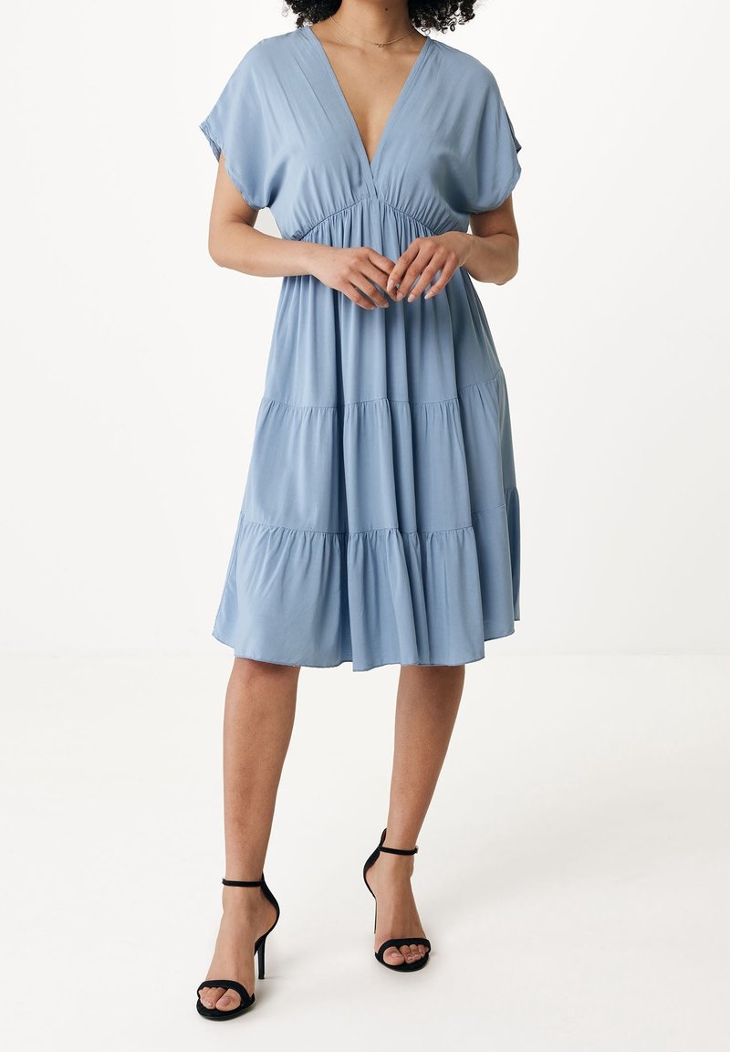 Svetle modré šaty do kolen s hlubokým V-výstřihem, krátkými rukávy a vrstvenou sukní. Vyrobené z měkké tkaniny s hladkým povrchem. Černé sandály na otevřenou špičku.