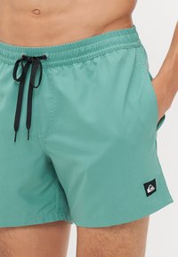 Lys tealfargede svømmebukser laget av hurtig tørkende materiale, med svart snor, sidelommer og en liten logo-patch på kanten.