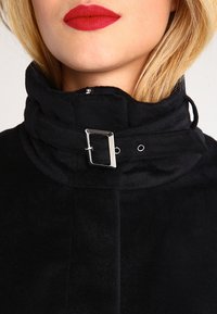 Giacca nera con colletto alto, dotata di una fibbia argentata, texture morbida e dettaglio di chiusura a pressione. Focalizzate su linee pulite e design moderno.