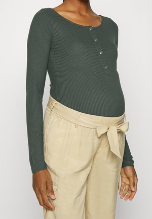 Personne enceinte portant un haut moulant côtelé vert foncé à manches longues avec des boutons et un pantalon beige taille haute noué avec un nœud.