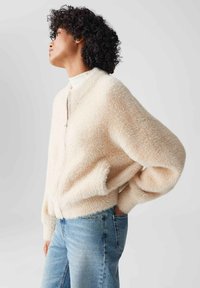 Frau mit lockigem Haar, die eine beiges, flauschiges Reißverschlussjacke und hellblaue Jeans trägt, steht mit einer Hand in der Tasche und blickt zur Seite.