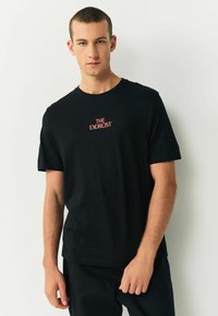 Melnas kokvilnas T-krekls ar īsām piedurknēm, ar sarkanu izšuvumu "THE EXORCIST" uz krūtīm. Vienkāršs apaļa kakla izgriezums, brīvs piegriezums.