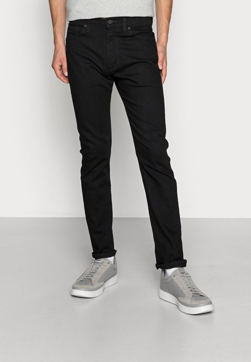 Homme portant un jean noir slim retroussé aux chevilles et des baskets décontractées gris et blanc, debout sur un sol clair contre un mur blanc.