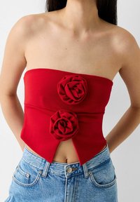 Rotes trägerloses Oberteil mit zwei Stoffakzenten in Blumenmuster, eng anliegendem Design und ausgeschnittenem Vorderteil. Kombiniert mit hochgeschnittenen Jeansshorts.