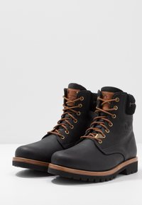 Botas de cuero negras con acentos marrones, punta redonda, ojales de metal y cordones marrones; superficie texturizada y suela de agarrado robusto.