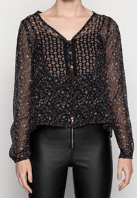 Svart genomskinlig blus med blommig och paisleytryck, V-ringning och knappslå. Långa ärmar med elastiska manschetter, parat med svarta byxor.