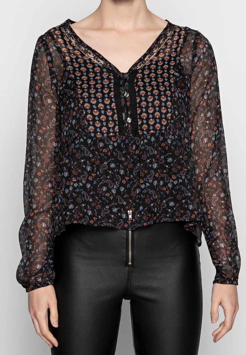 Svart genomskinlig blus med blommig och paisleytryck, V-ringning och knappslå. Långa ärmar med elastiska manschetter, parat med svarta byxor.