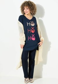 Marineblaues Tunika-Shirt mit "HO HO HO" in verschiedenen Farben bedruckt; kombiniert mit dunkelblauen Jeans und einer cremfarbenen, offen gestrickten Strickjacke. Metallische Schuhe runden den Look ab.