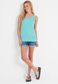Woman standing against white wall, wearing turquoise sleeveless top, rolled-up denim shorts, and metallic buckle sandals, smiling and looking down.