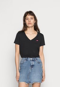 Černé tričko s výstřihem do V a krátkými rukávy, na kterém je malé červené logo Levi's, zkombinované s modrou džínovou sukní s roztrhaným lemem.