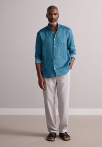 Camisa de lino turquesa con un patrón sutil, pantalones beige claro y sandalias negras. La camisa tiene un cuello abotonado y las mangas remangadas.